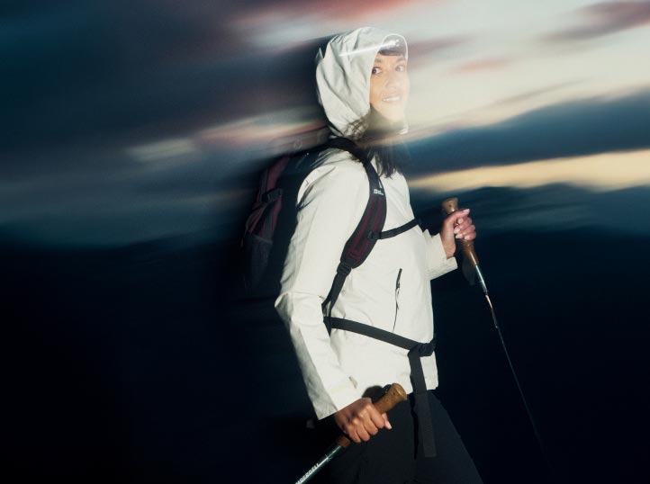 Una persona con ropa de senderismo y chaqueta blanca con capucha sonríe mientras sujeta sus bastones de senderismo. El cielo crepuscular difuminado sugiere movimiento y aventura.
