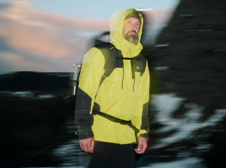 Un hombre con chaqueta amarilla brillante y mochila haciendo senderismo al atardecer, el desenfoque del movimiento le da dinamismo a la imagen.