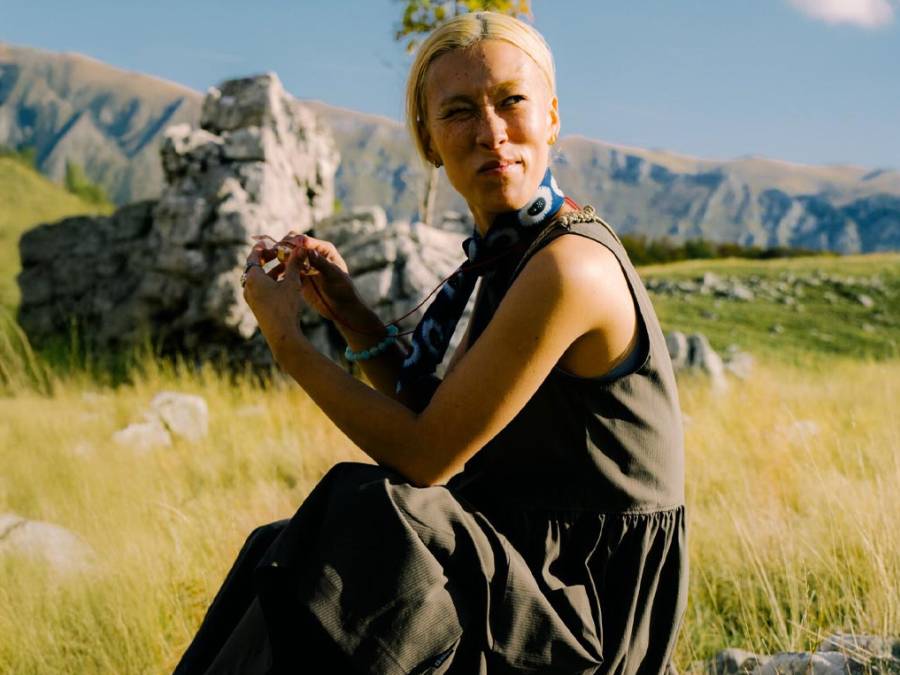 Una mujer rubia sentada en una campa verde, mira a lo lejos; está rodeada de montañas.