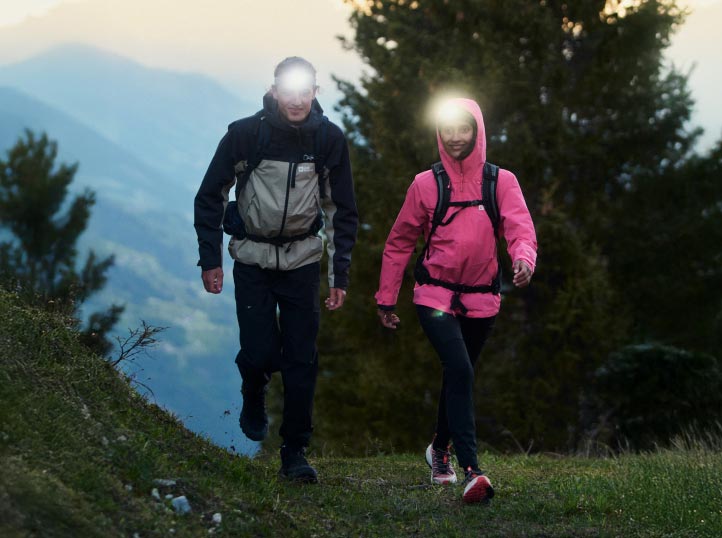 Dos senderistas con linternas frontales en un sendero forestal mientras anochece. Llevan chaquetas, una es negra y gris y la otra rosa.